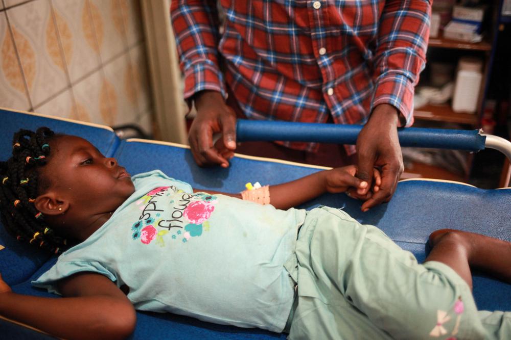 Collins consuela a su hija, y le habla de la importancia de ser vacunada para tener buena salud, para distraerla del trozo de cristal que le están quitando del pie. Bamenda, noroeste de Camerún.